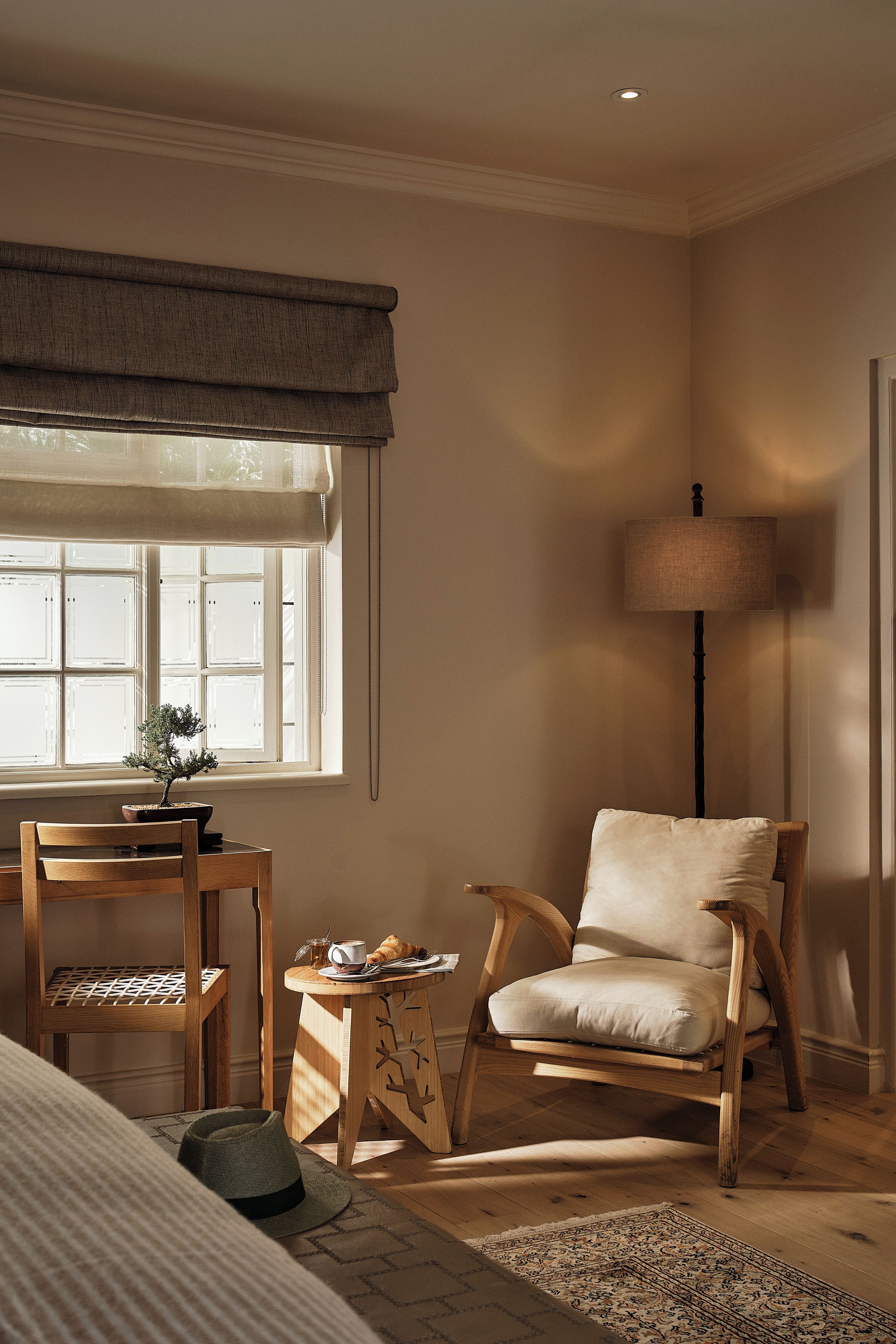Una habitación con una silla de madera, una mesita con una planta, una lámpara y una ventana con persianas.