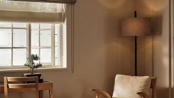 Una habitación con una silla de madera, una mesita con una planta, una lámpara y una ventana con persianas.