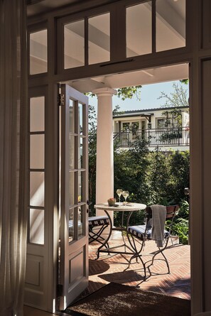 An open door leading to a patio with a table and chairs, a view of a building across the street, and a hedge in the background. - Leeu House (Franschhoek)