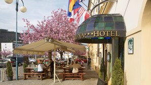 Ein Sitzbereich im Freien mit Tischen und Bänken unter einer überdachten Terrasse, ein Schild mit der Aufschrift "Kuna Hotel" und Fahnen an einem Gebäude.