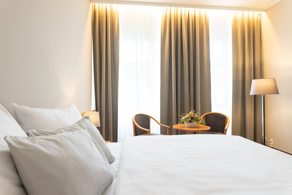 A hotel room with a bed, two chairs, a small table with a flower vase, and curtains.