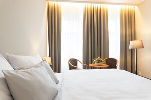 Una habitación de hotel con una cama, dos sillas, una mesita con un florero y cortinas.