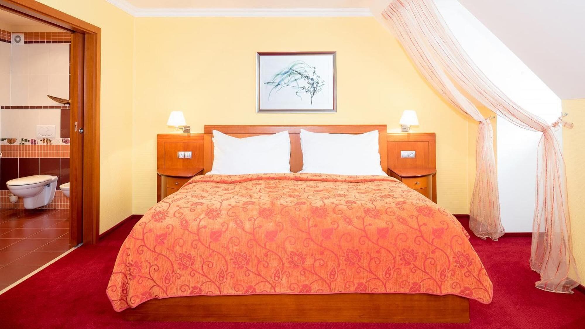 Habitación de hotel con una cama, dos mesitas de noche, un tocador, un cuadro enmarcado en la pared y un baño visible a través de una puerta abierta.