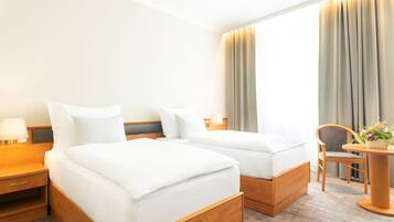 A hotel room with two single beds, a wooden nightstand with a lamp, a small table with a plant, and a chair.