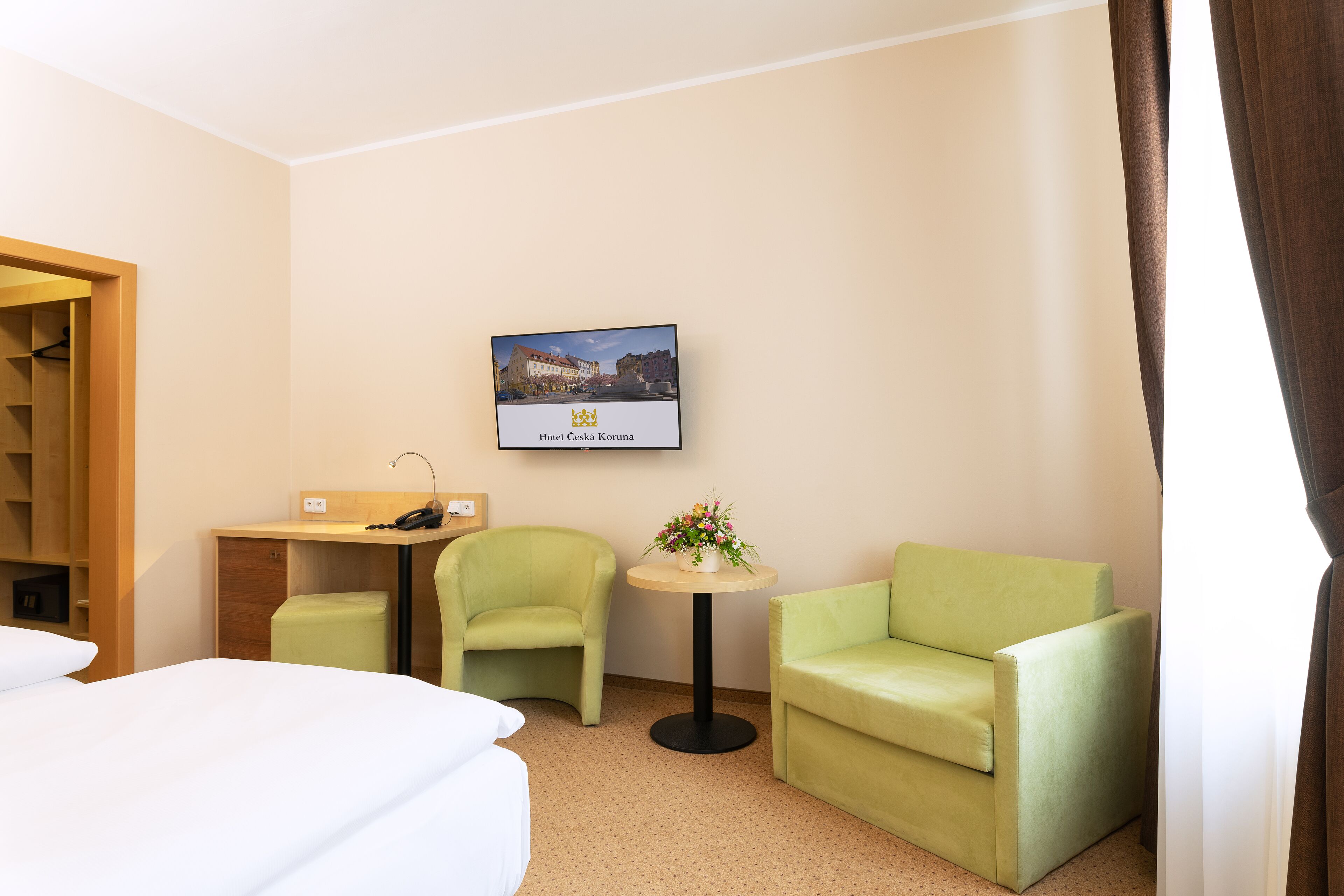 A hotel room with a bed, two green armchairs, a small table with a flower vase, a desk with a phone, and a framed picture of a building.
