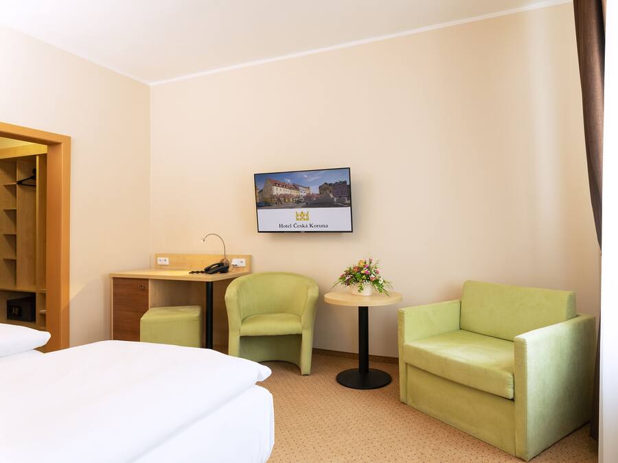 Una habitación de hotel con una cama, dos sillones verdes, una mesita con un florero, un escritorio con un teléfono y un cuadro con la imagen de un edificio.