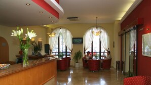 Eine Hotelhalle mit Empfangstresen, einer Sitzecke und großen Fenstern mit Vorhängen.