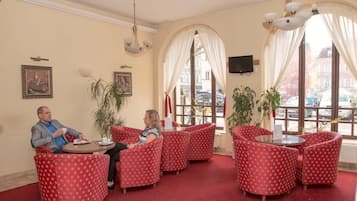 A hotel lobby with red patterned chairs, a round table, a large window with curtains, and a television mounted on the wall.