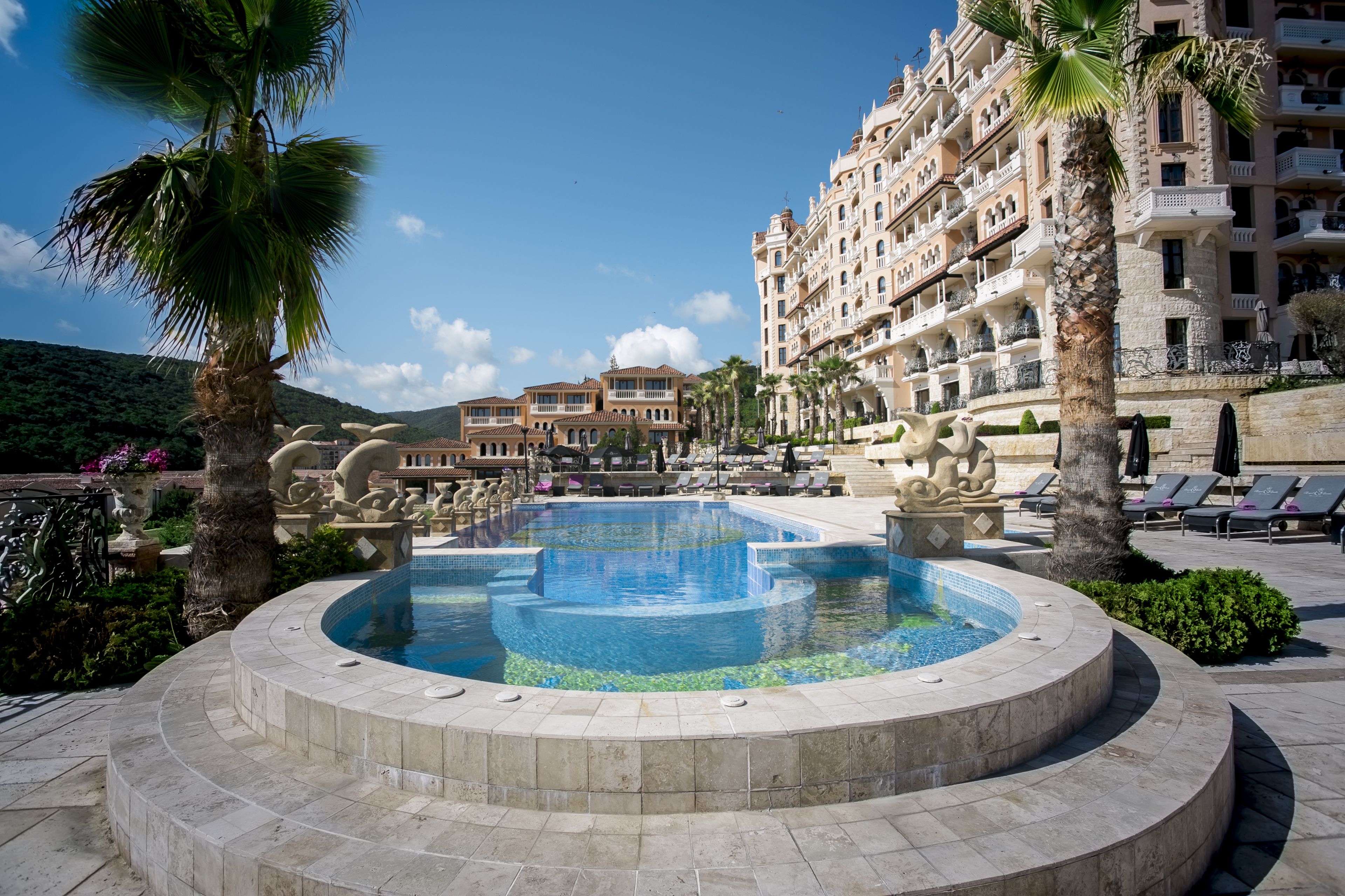A pool area with a circular design, surrounded by palm trees and lounge chairs, with a large building in the background.