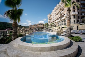 Un área de piscina con diseño circular, rodeada de palmeras y sillones de descanso, con un edificio grande al fondo.