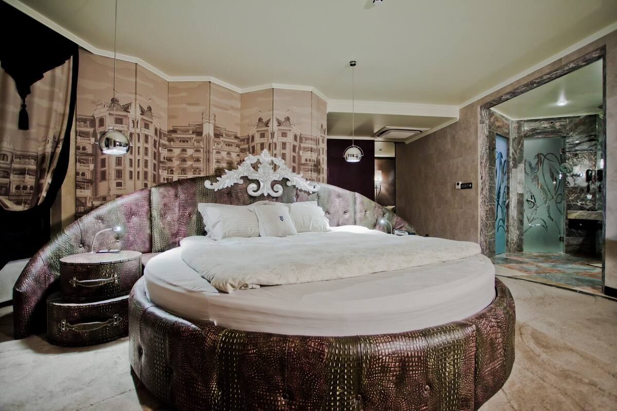 A bedroom with a large bed, a round ottoman, a mirror, and a wall with a cityscape mural.