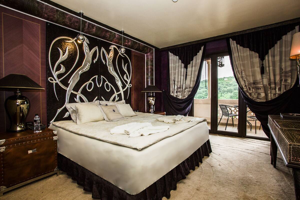A large bed with a decorative headboard, a bedside table with a lamp, and a view of greenery through the window.