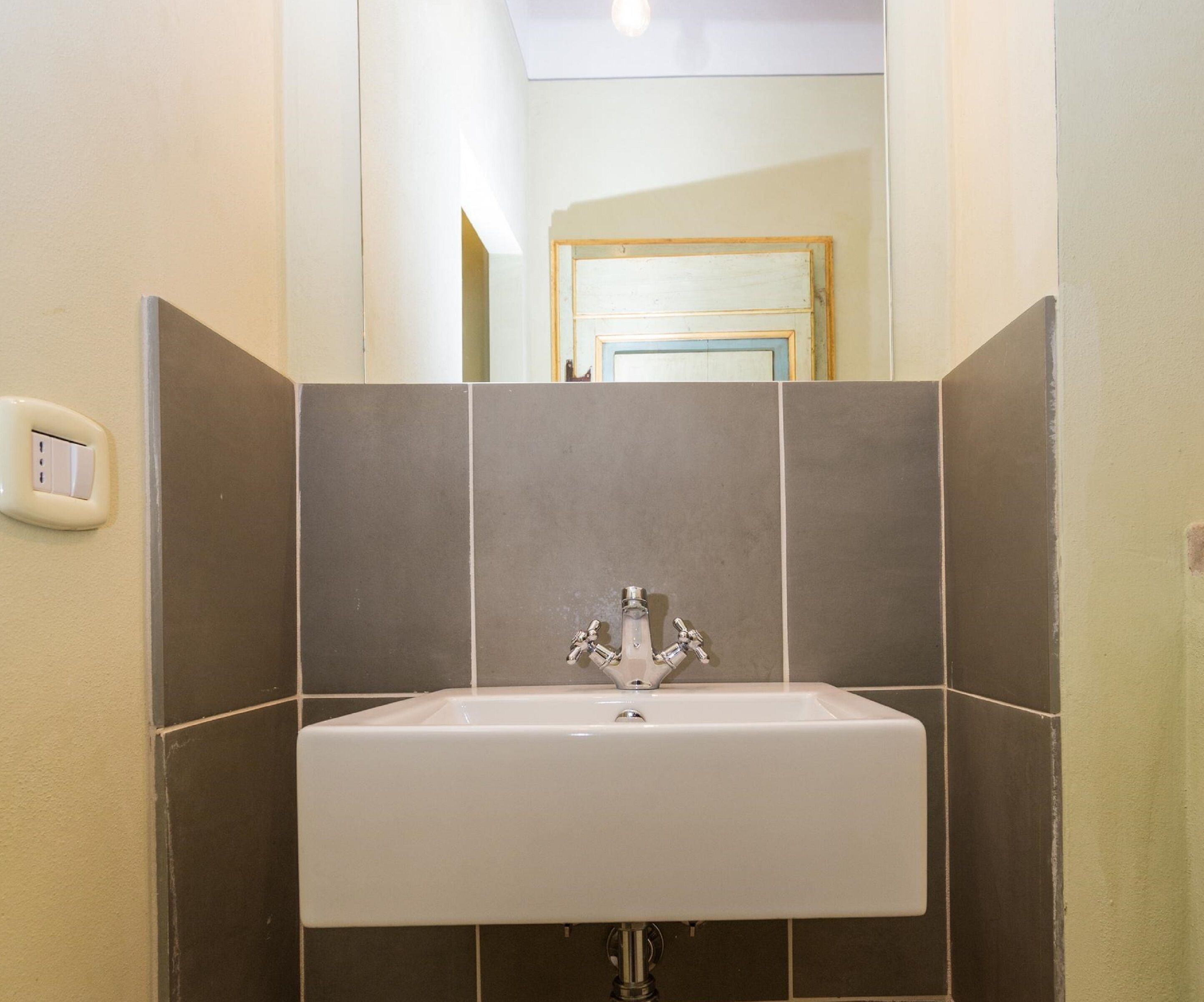 A bathroom with a white sink, dark tiled walls, and a visible light switch on the left.
