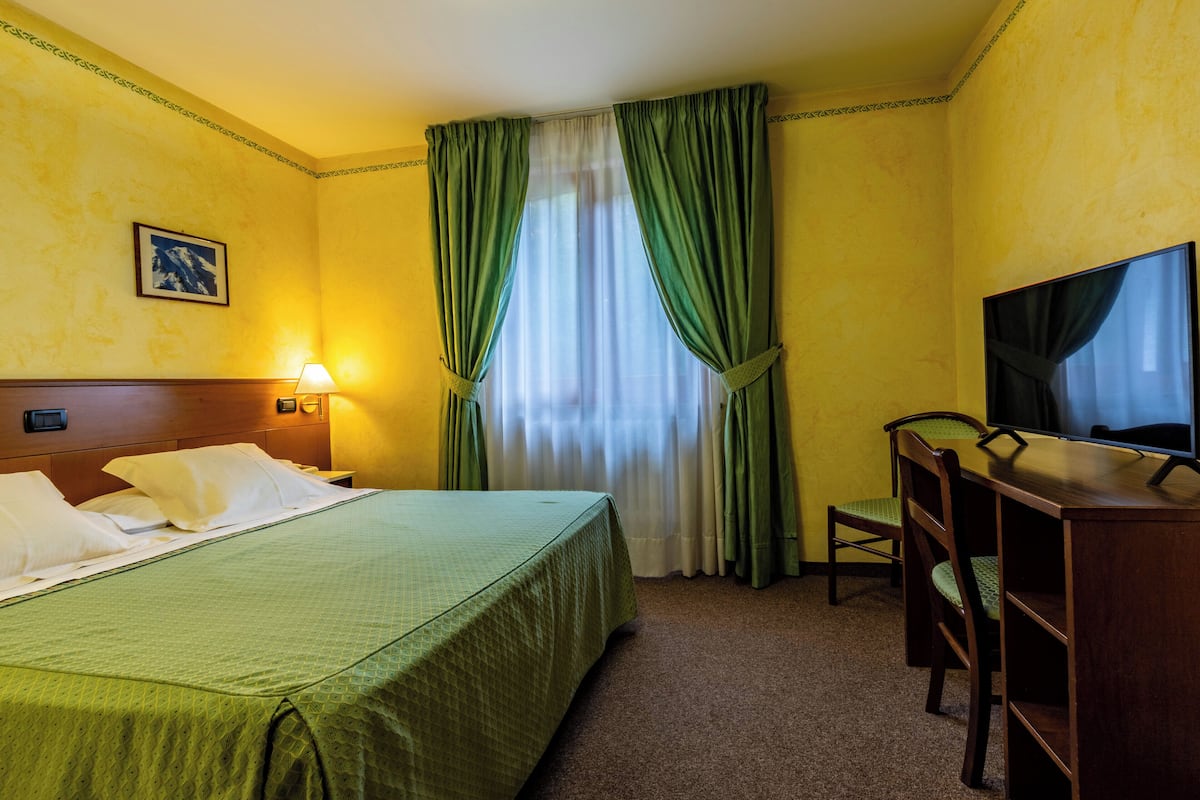 Habitación de hotel con cama, escritorio, silla, televisor y ventana con cortinas.