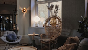 A cozy seating area with a wicker chair, a grey armchair, and a small round table with a candle. - Home Hotel Borgen (Örebro)
