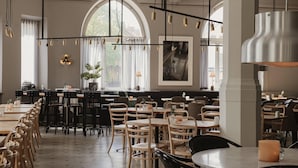 A restaurant with high tables and chairs, hanging pendant lights, and a large arched window. - Home Hotel Borgen (Örebro)
