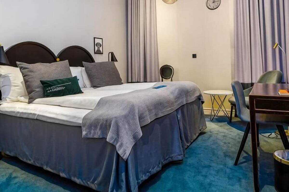 A neatly made bed with a grey bedspread and pillows, a small round table with a chair, and a blue carpeted floor.