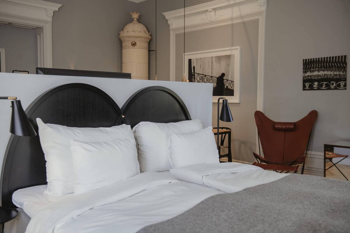 A bed with a black headboard, white bedding, a brown leather chair, a small table with a lamp, and a decorative wall piece.