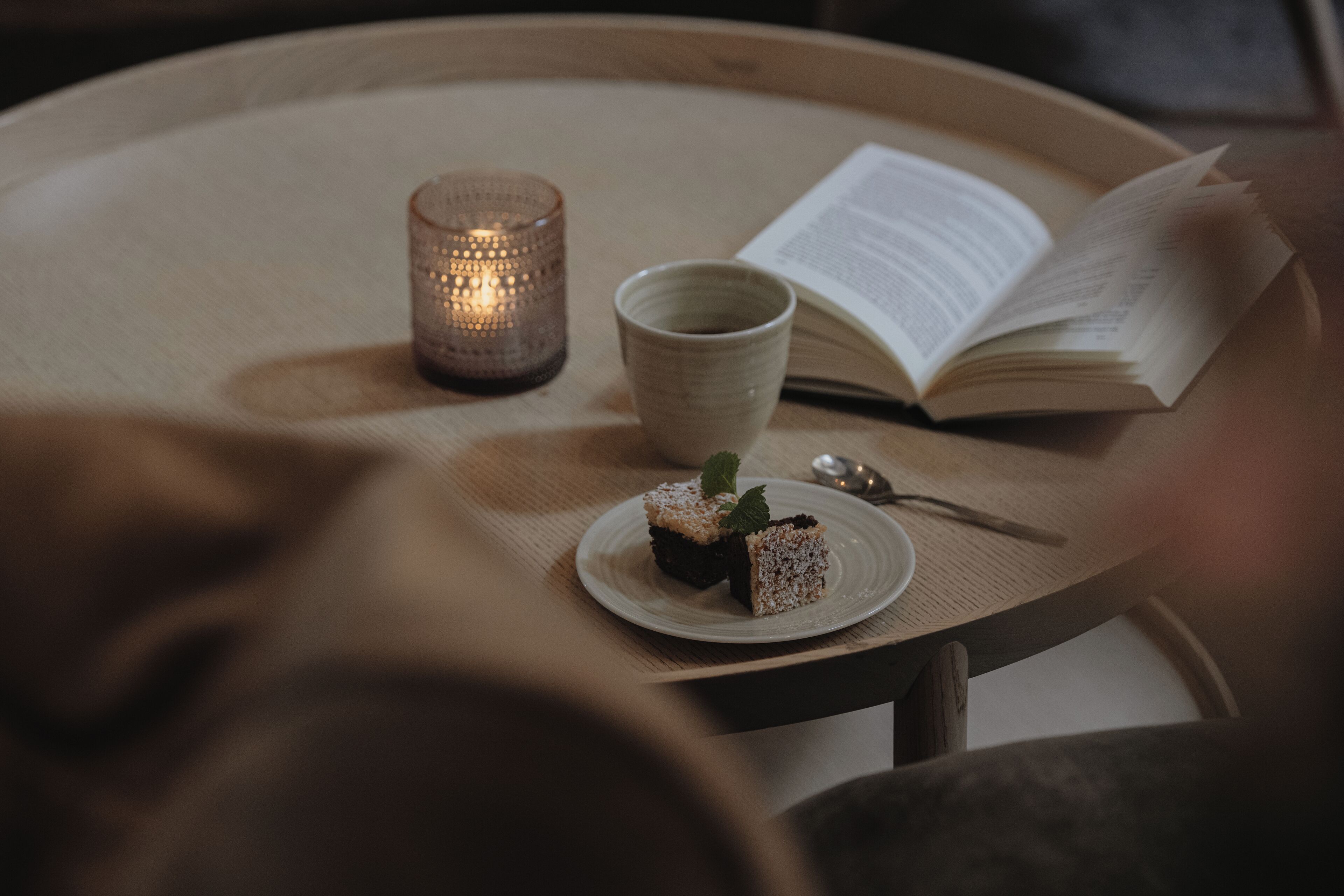 A round wooden table with a candle, a cup, a book, and a plate with a dessert.