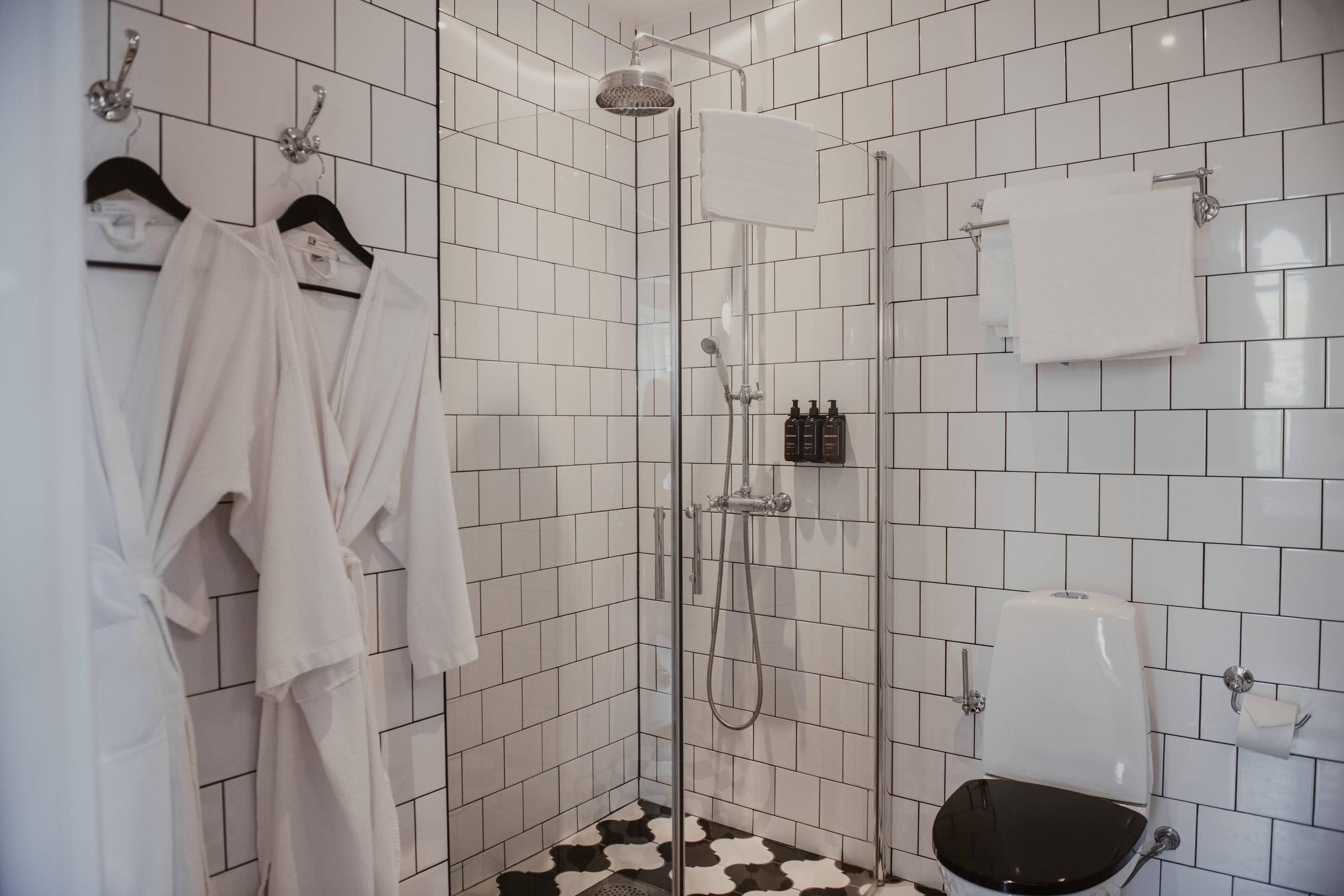 Kylpyhuoneen seinät ovat valkoisilla laatoilla, siinä on lasiovilla varustettu suihku, wc ja kaksi valkoista kylpytakia, jotka roikkuvat henkaritelineessä.