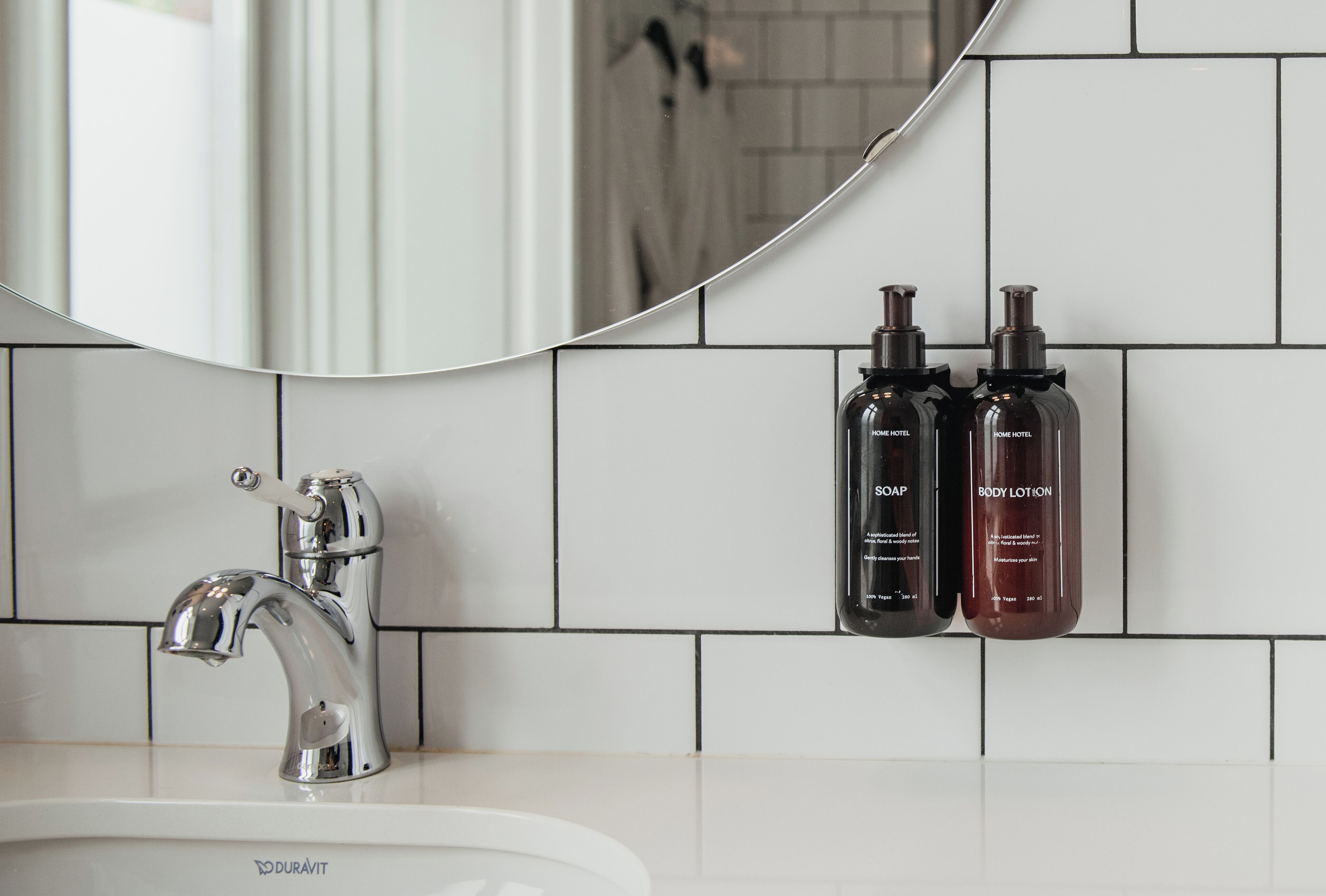 Ein Badezimmer mit einem weißen Waschbecken, einem Chrom-Wasserhahn und zwei schwarzen Flaschen mit den Aufschriften "SOAP" und "BODY LOTION" an der Wand.