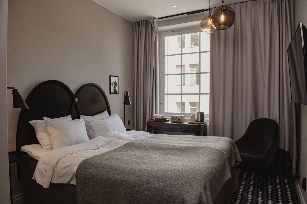 A hotel room with a large bed, a chair, a desk with a radio, and a window with curtains.