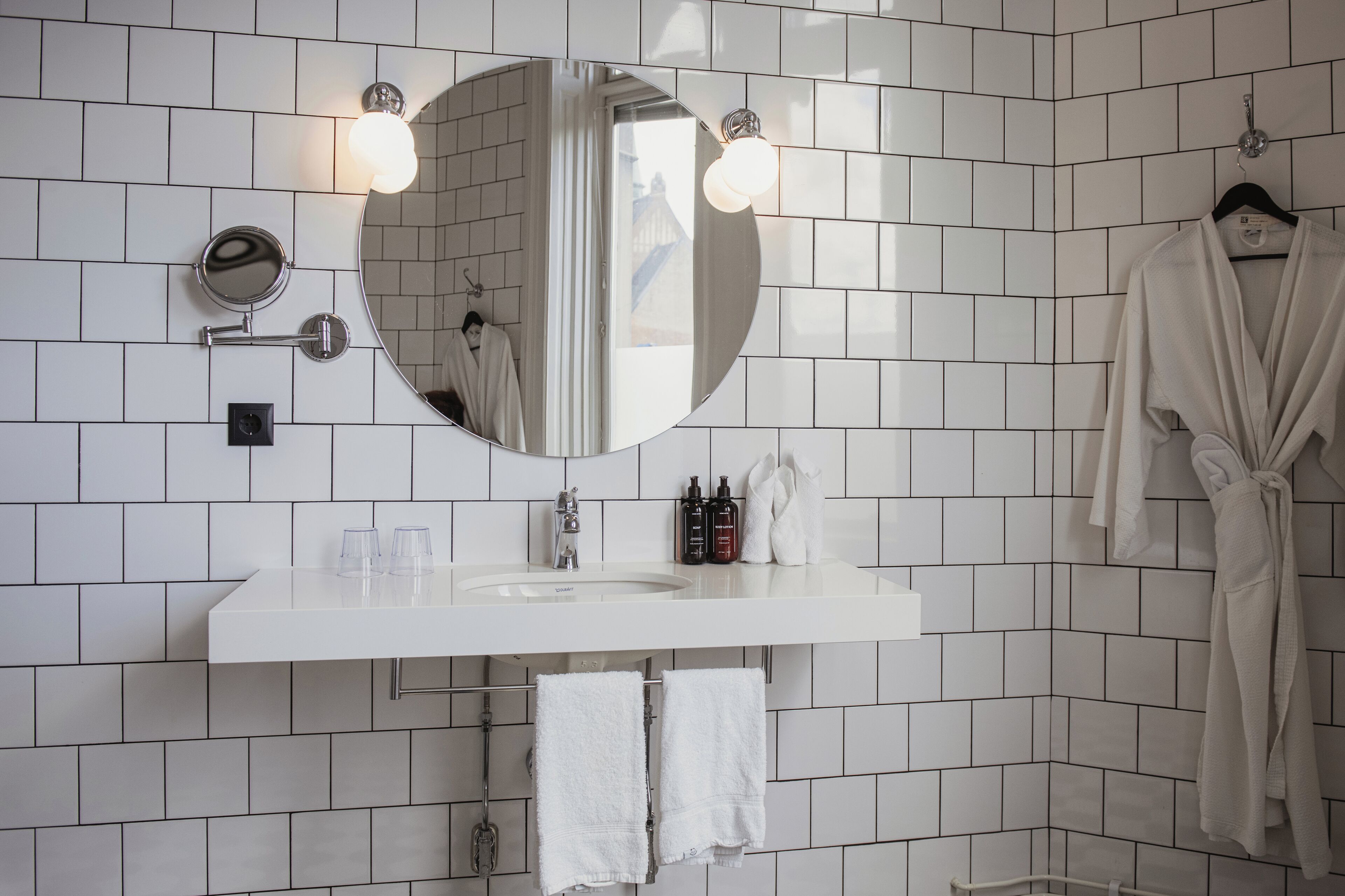 Un baño moderno con pared de azulejos blancos, un espejo redondo con iluminación, un lavabo blanco y una bata colgada en la pared.