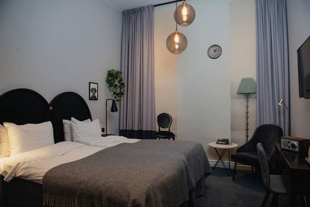 A hotel room with a bed, a desk, a chair, a lamp, curtains, a clock, and a plant.