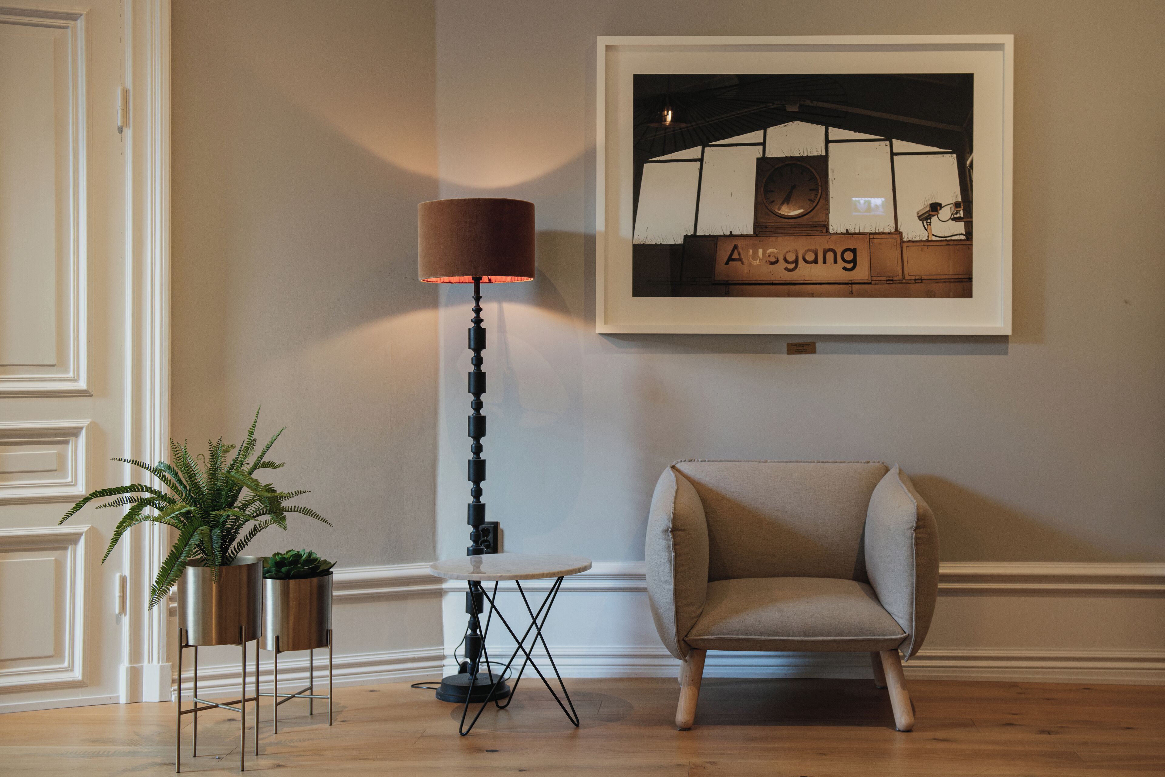 A room with a framed picture of a bar, a floor lamp, a chair, and two potted plants.
