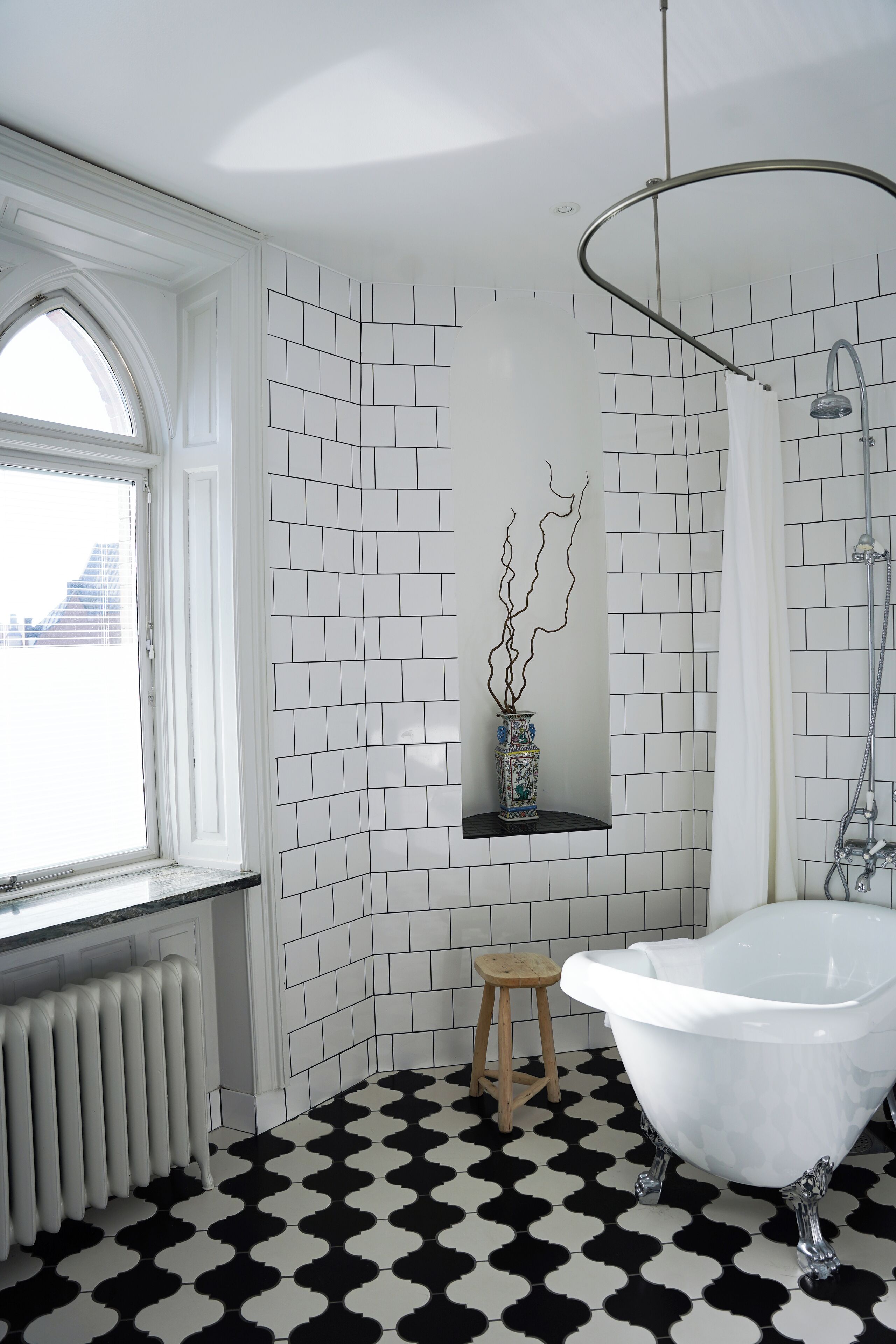 Un baño con una bañera blanca, un taburete de madera, un radiador y una ventana con vistas a edificios.