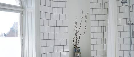 Un baño con una bañera blanca, un taburete de madera, un radiador y una ventana con vistas a edificios.