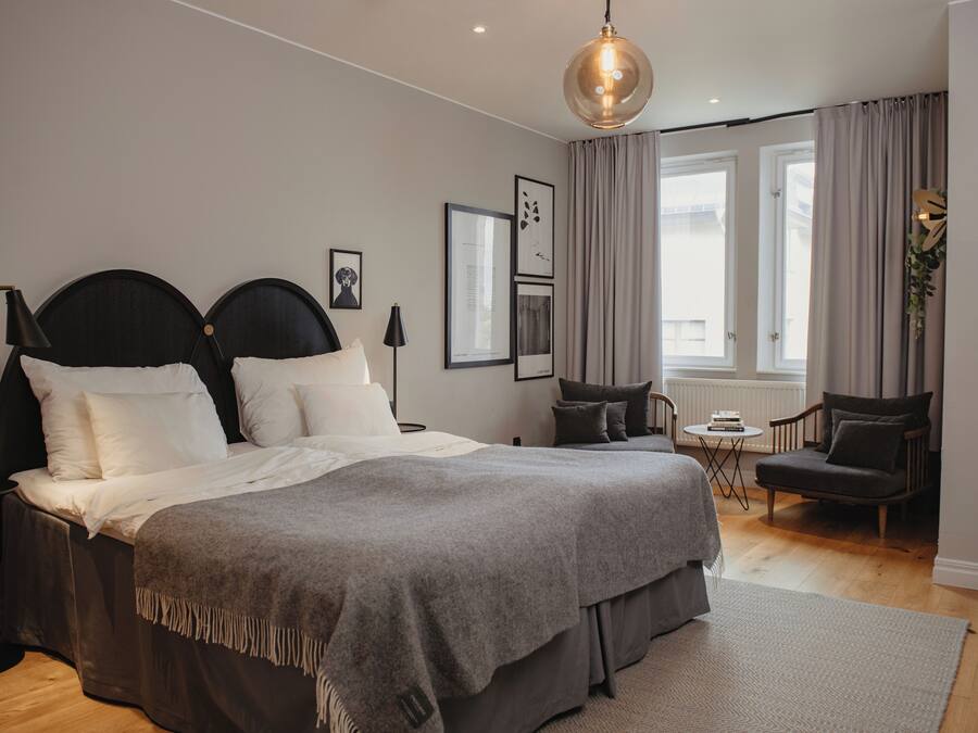 A modern bedroom with a large bed, a small side table, a chair, and a view of the outdoors through large windows.