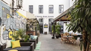 Een binnenplaats met een zwarte bank, gele kussens en een gestreept vloerkleed. Er staan potplanten en een tafel met stoelen. Op de linker muur is een muurschildering te zien, en de naam van het hotel is zichtbaar boven de ingang.