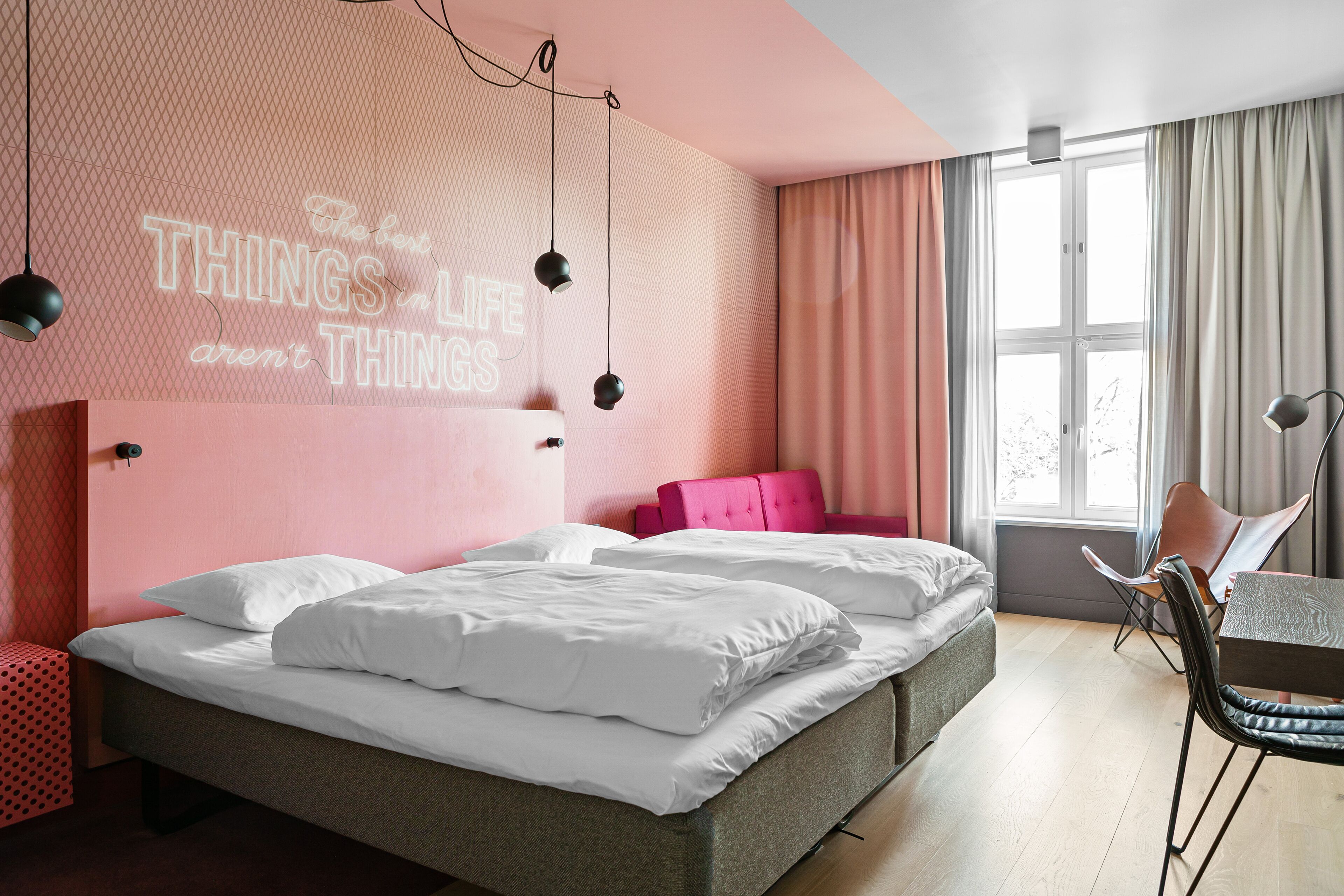 Un dormitorio moderno con cama, pared rosa con una cita motivacional, ventana con cortinas y una silla.