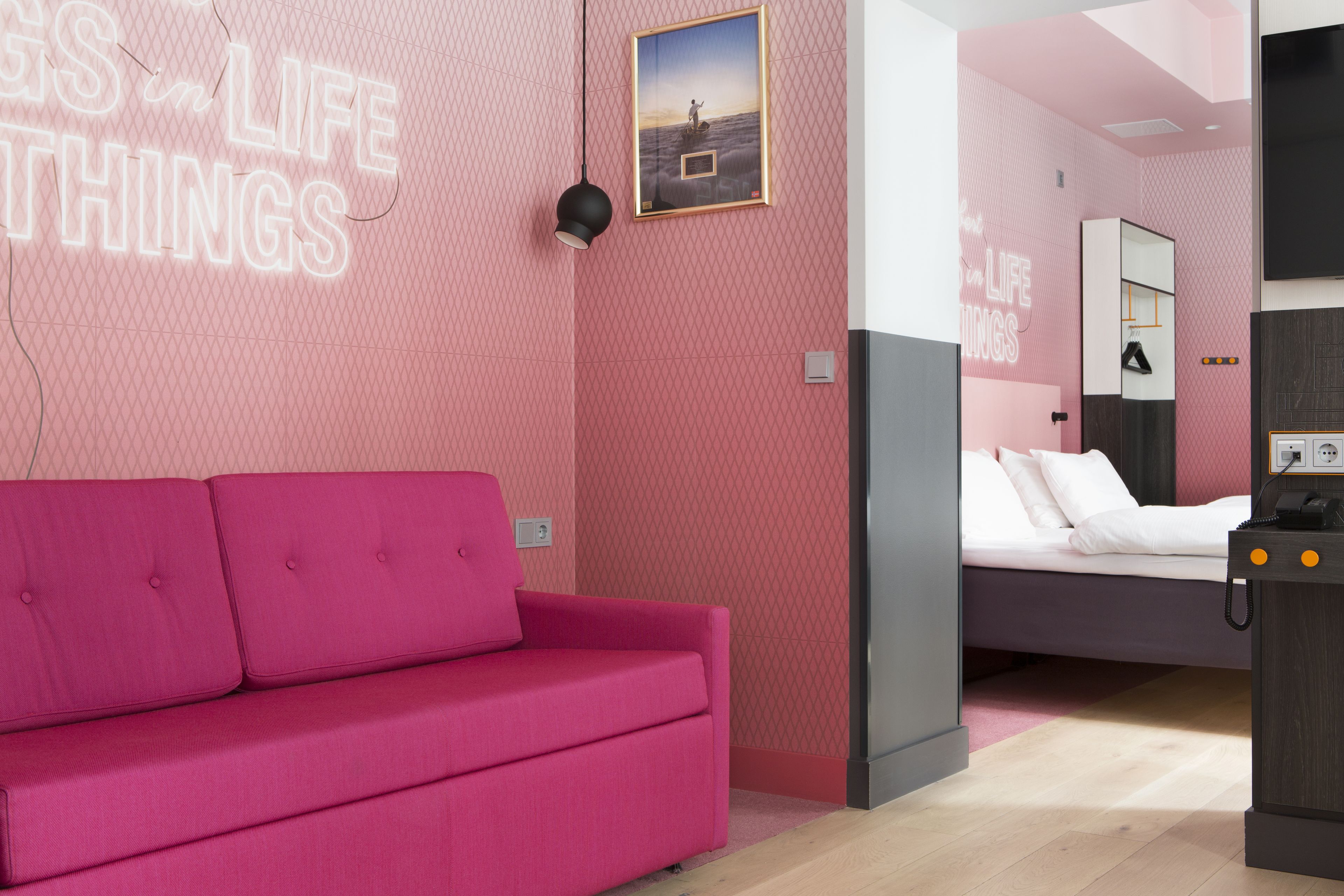 A modern hotel room with a pink wall, a bed, a desk with a phone, and a neon sign.