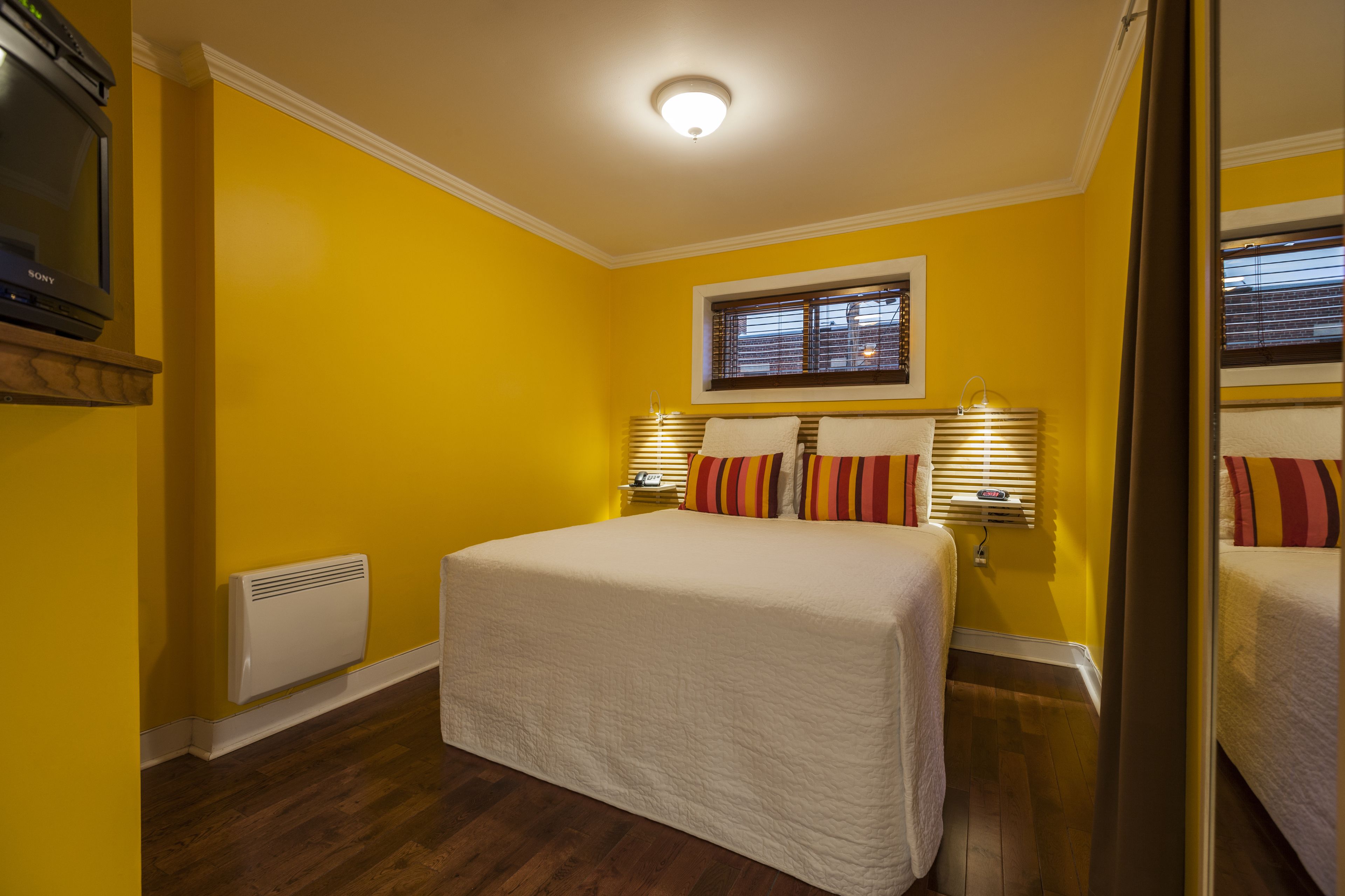 A room with a large bed, two bedside lamps, a window with blinds, a wall-mounted heater, and a television on a shelf.