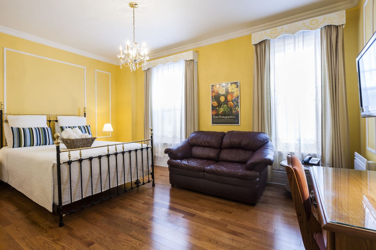 Habitación de hotel con cama, sofá de cuero marrón, mesa de comedor de madera, candelabro y ventana con cortinas.