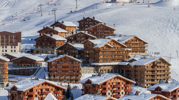 Un conjunto de cabañas de madera en la montaña con varios balcones, situadas en una ladera nevada con teleféricos al fondo.