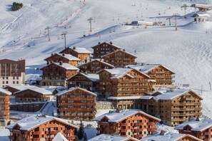 Un ensemble de chalets de montagne en bois, dotés de plusieurs balcons, disposés sur une pente enneigée, avec des remontées mécaniques en arrière-plan.