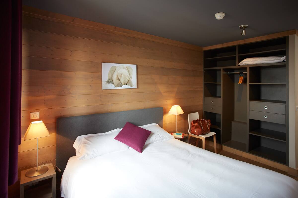 Una habitación de hotel con cama, dos lámparas de noche, mesita de noche, cabecero de madera, perchero y armario.
