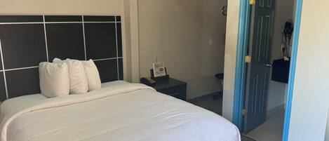 Un lit bien fait, avec une tête de lit, une table de chevet sur laquelle se trouvent un téléphone et un petit objet, et une porte qui donne sur une autre pièce.