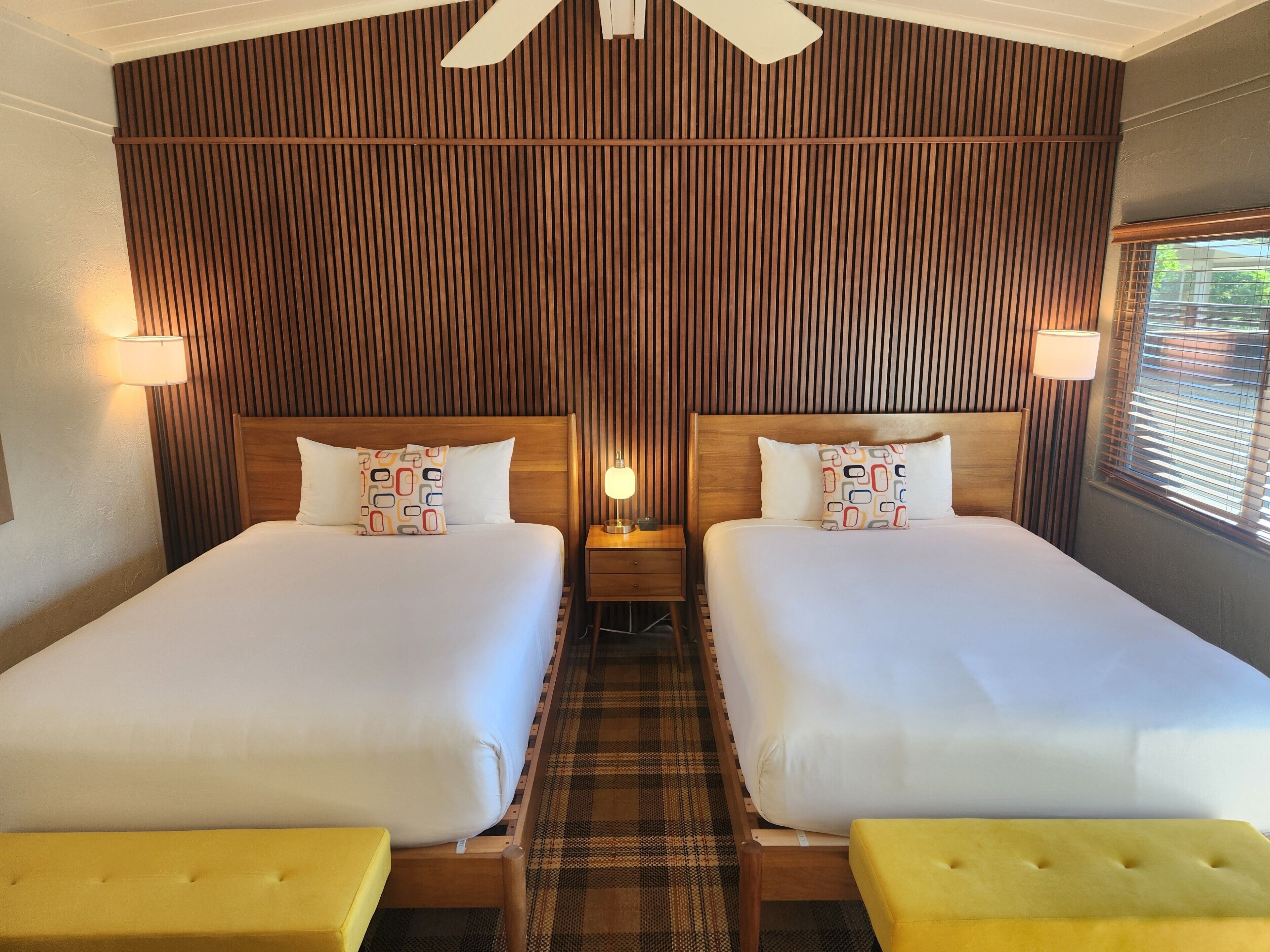 A hotel room with two beds, a wooden panel wall, a ceiling fan, and a window with blinds.