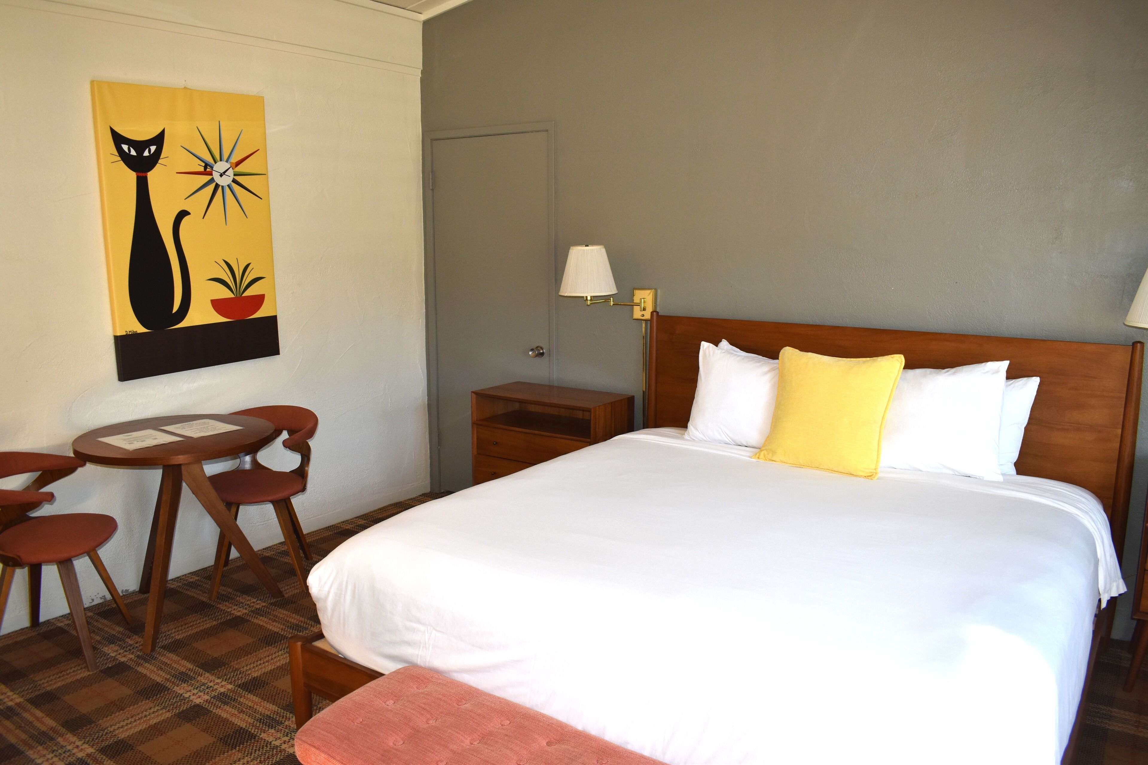 A hotel room with a large bed, a small table with chairs, a nightstand, a lamp, and a wall painting of a cat.