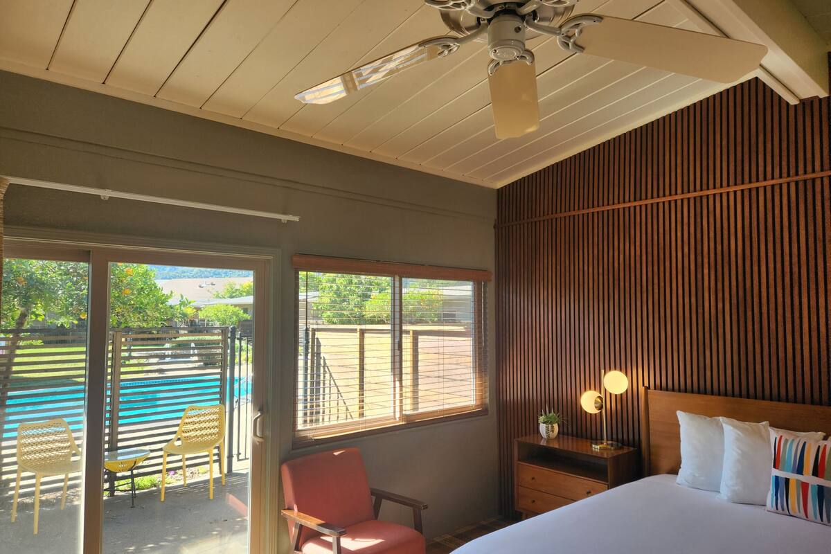 Un dormitorio con cama, mesita de noche, silla, ventana con persianas, ventilador de techo y vista a un área de piscina al aire libre.