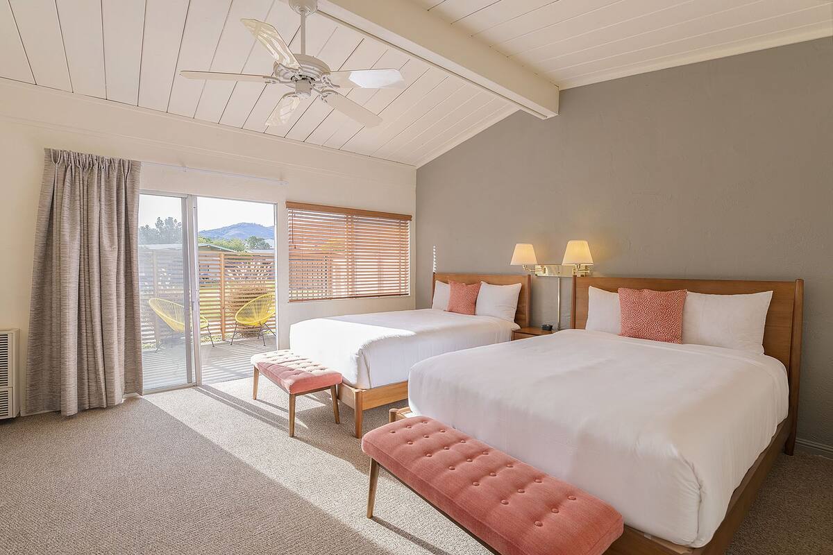 Habitación de hotel con dos camas, ventilador de techo, balcón con vistas y una banca.