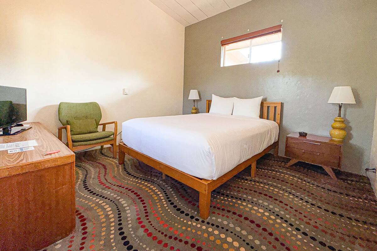 Un dormitorio con una cama de madera, una silla verde, un escritorio con una computadora y una alfombra estampada.