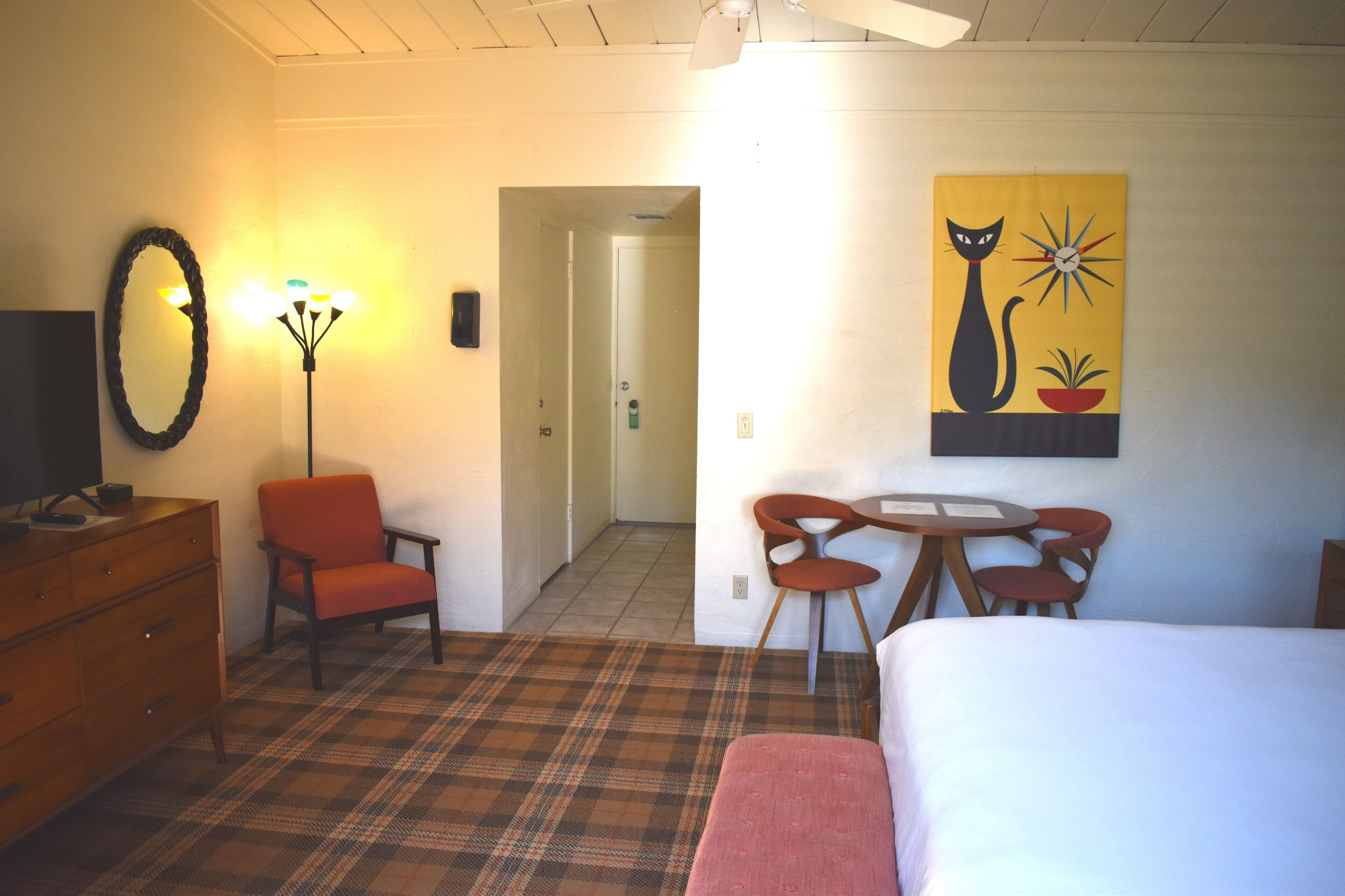 Una habitación de hotel con una cama, un escritorio con dos sillas, un espejo, una lámpara y un cuadro de un gato.