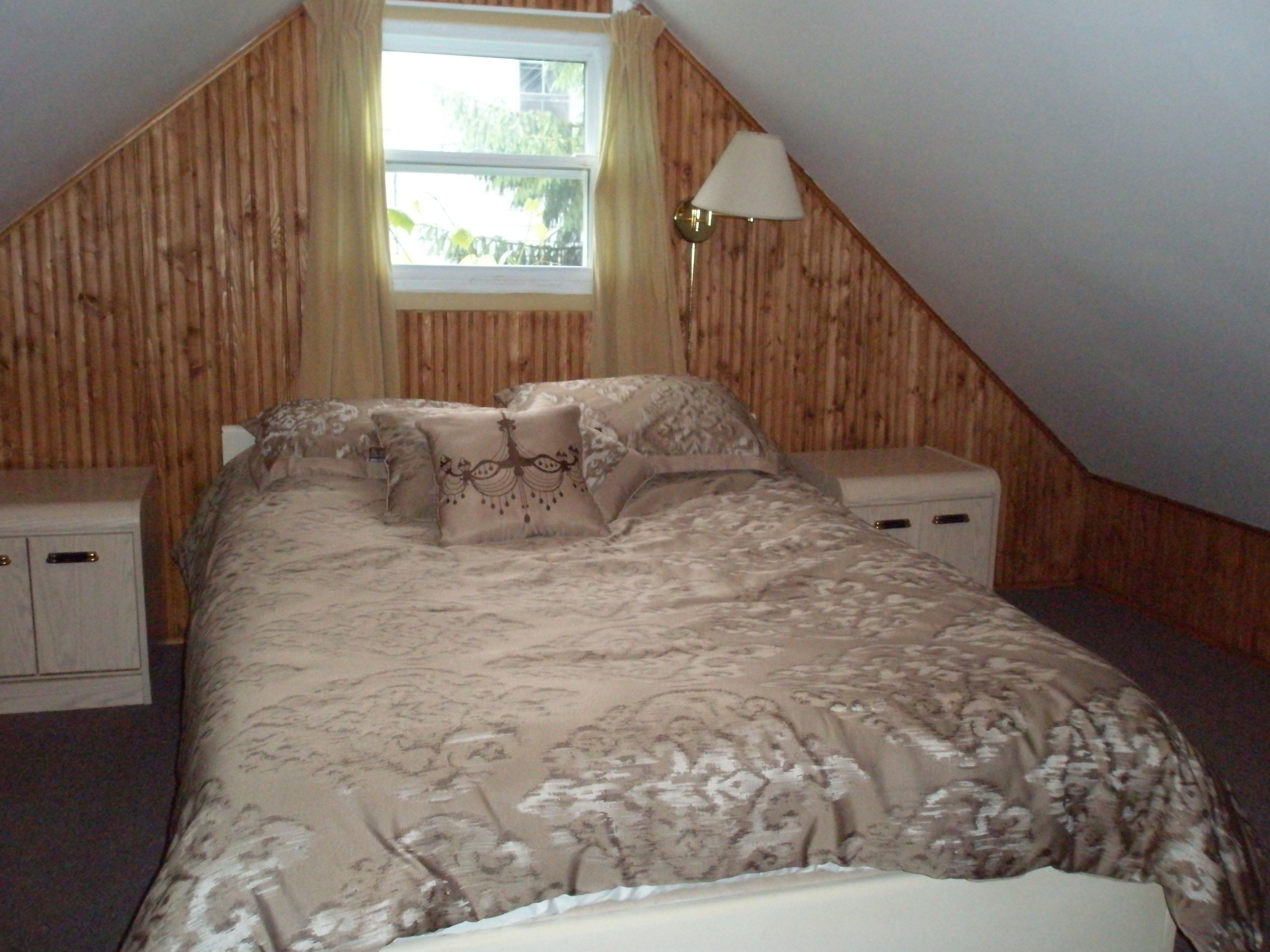 A bedroom with a sloped ceiling, a bed with floral bedding, two bedside tables, a lamp, and a window with curtains.