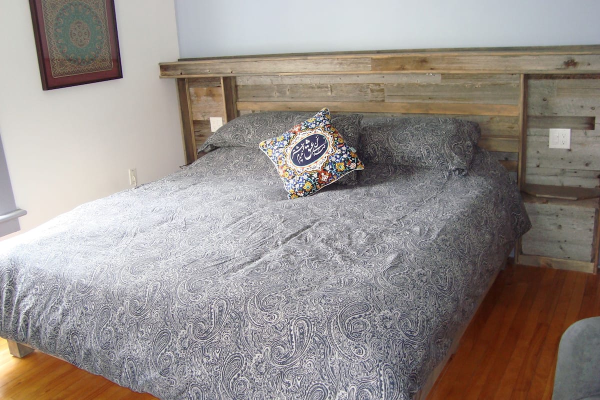 A bed with a patterned bedspread and a decorative pillow, a wooden headboard, and a wall-mounted picture frame.