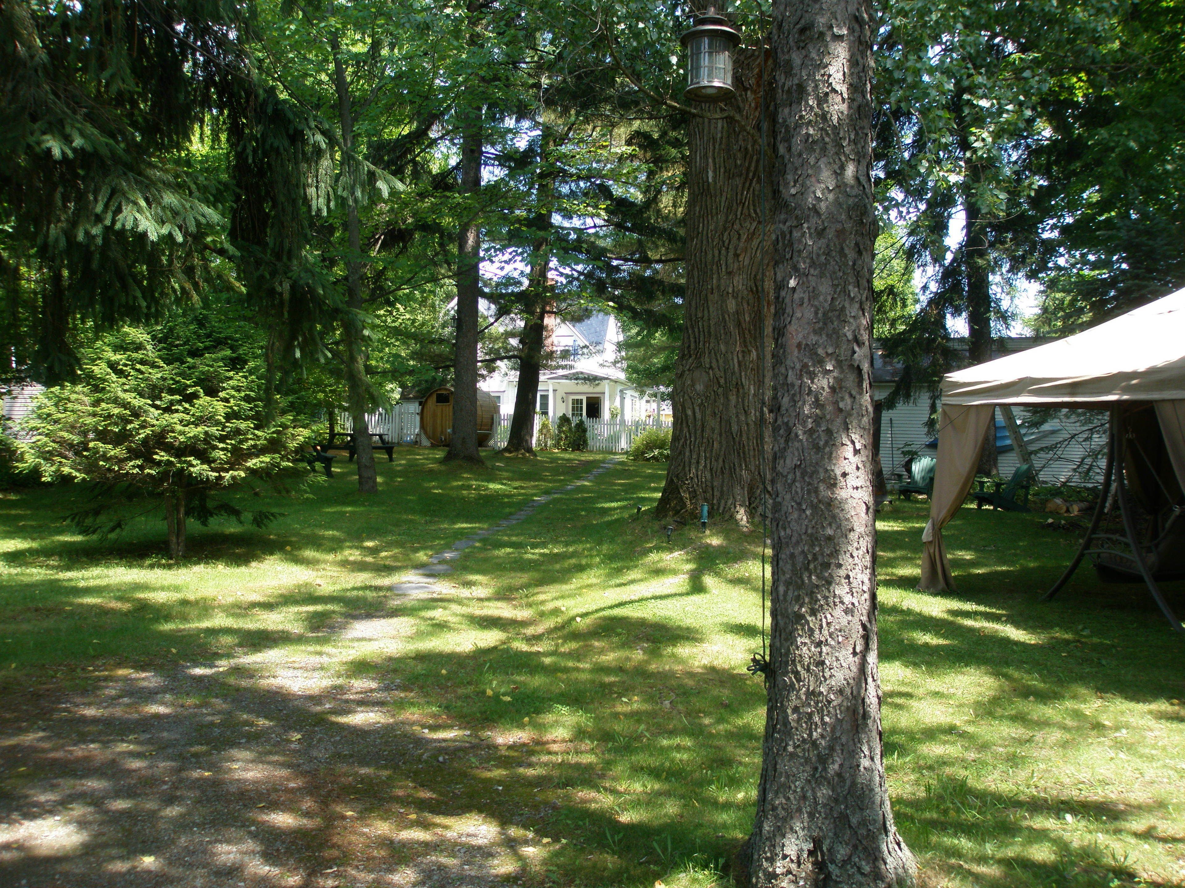 A forested yard with a path leading to a white house, a wooden barrel, and a tent.